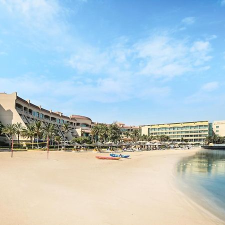 Al Raha Beach Resort & Spa Abu Dhabi Bagian luar foto The photo depicts a serene beach scene with a clear blue sky. In the foreground, there is a sandy beach that gently slopes down to calm water. On the left side, several palm trees provide a tropical atmosphere. In the background, there are two large 