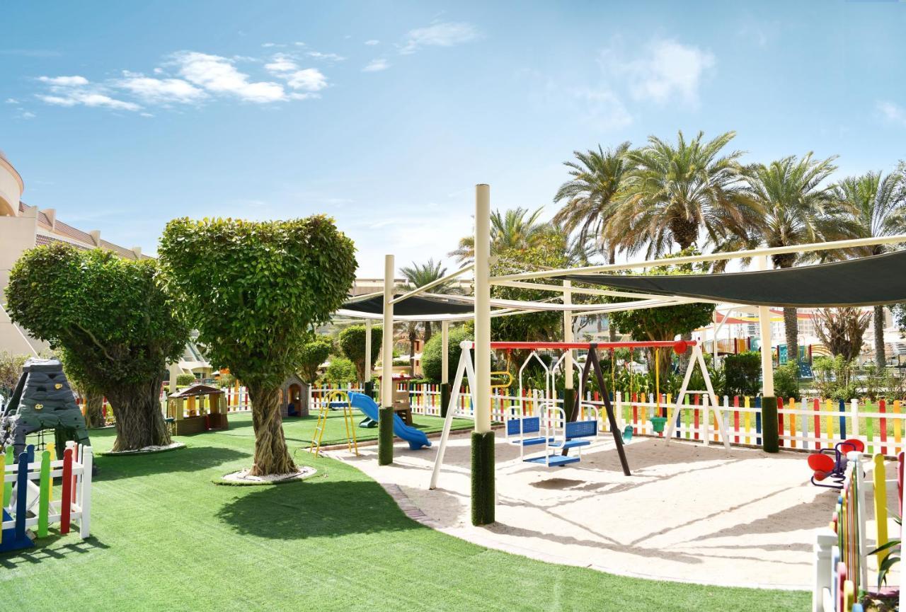 Al Raha Beach Resort & Spa Abu Dhabi Bagian luar foto The photo shows a brightly lit outdoor playground surrounded by palm trees. The playground features a variety of play equipment, including swings, a slide, and a climbing structure. The ground is covered in lush green artificial grass, providing a so