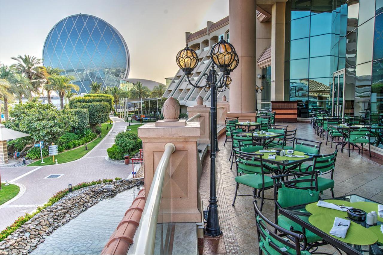 Al Raha Beach Resort & Spa Abu Dhabi Bagian luar foto The photo depicts an outdoor dining area with green tables and chairs arranged on a patio. In the background, there is a large, distinctive building with a striking blue dome, surrounded by well-maintained landscaping, including trees and plants. The