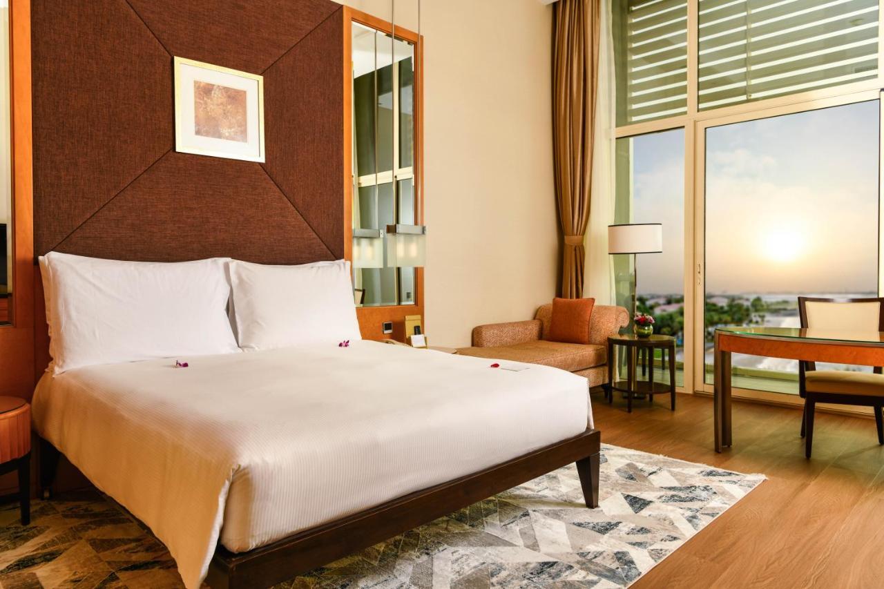 Al Raha Beach Resort & Spa Abu Dhabi Bagian luar foto The photo shows a stylish hotel room featuring a large bed with white bedding, positioned against a textured wall. There’s a small seating area with a chair and a table, and a window with curtains that allows natural light to fill the space. The room
