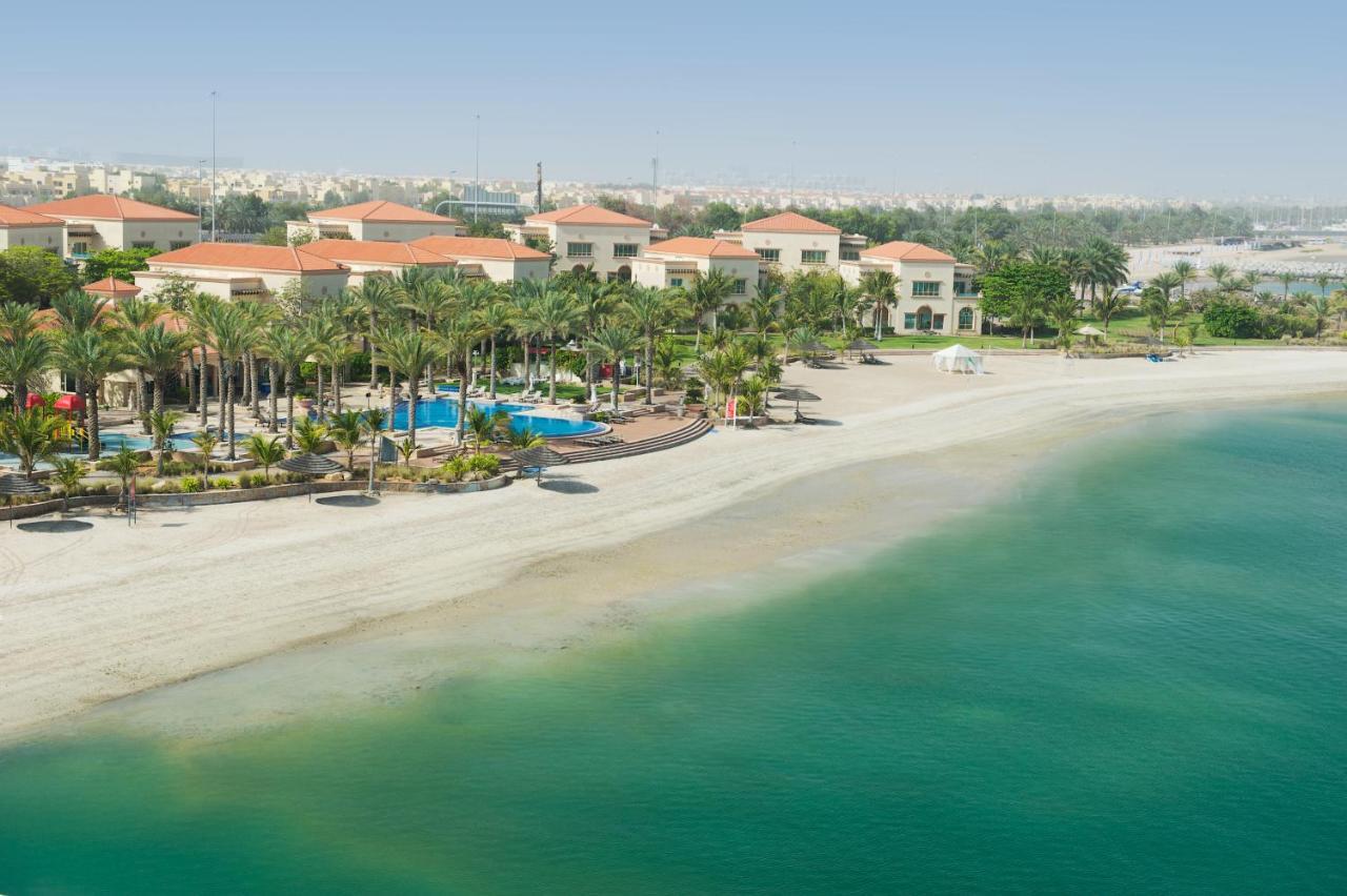 Al Raha Beach Resort & Spa Abu Dhabi Bagian luar foto The photo depicts a serene beach scene with a clear turquoise sea and a sandy shoreline. In the background, there are several buildings featuring a Mediterranean architectural style with red-tiled roofs. The area is adorned with palm trees and outdoo