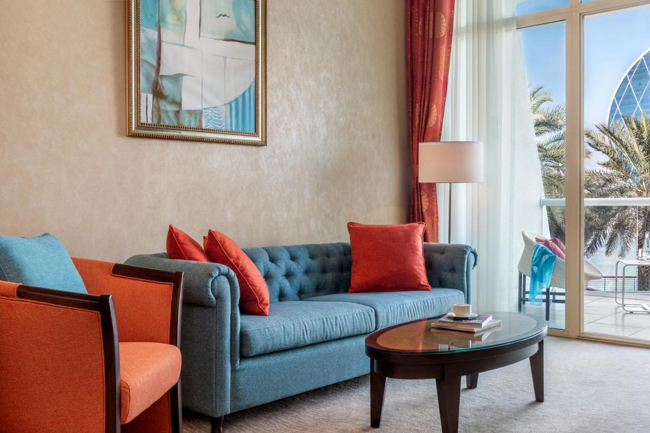 Al Raha Beach Resort & Spa Abu Dhabi Bagian luar foto The photo shows a well-decorated living room. There is a plush blue couch with orange accent pillows and a cozy orange armchair nearby. A round coffee table sits in front of the couch, and above it, there is a colorful painting hanging on the wall. L