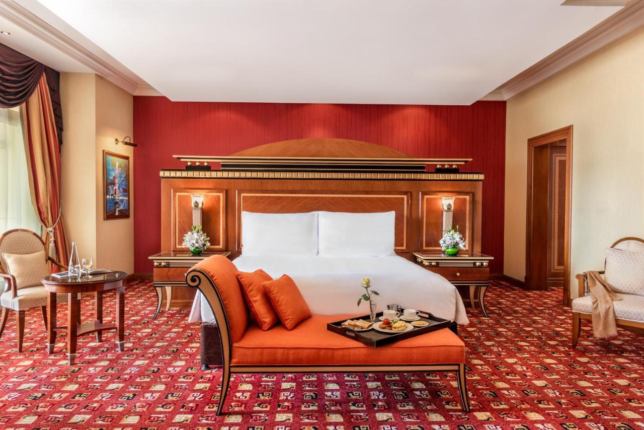 Al Raha Beach Resort & Spa Abu Dhabi Bagian luar foto The photo shows an elegantly designed hotel room. The room features a large bed with white linens and a decorative headboard. The walls are painted in a warm red color, and there are side tables with lamps beside the bed. 

There is an orange sofa or