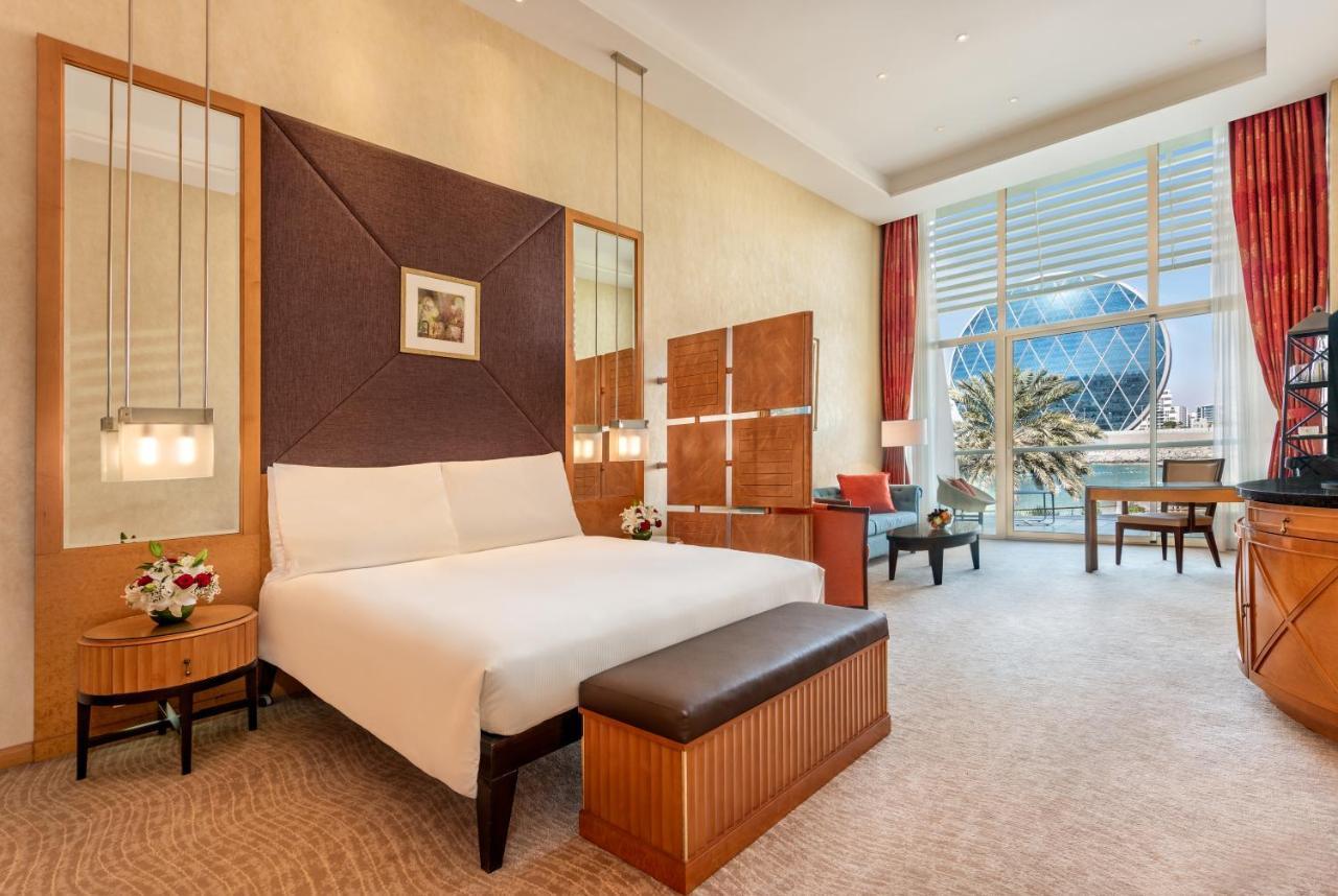 Al Raha Beach Resort & Spa Abu Dhabi Bagian luar foto The photo shows a modern hotel room featuring a neatly made bed with a white comforter, situated against a stylish headboard with a geometric design. There are bedside tables on either side of the bed, each with a small flower arrangement. A comforta