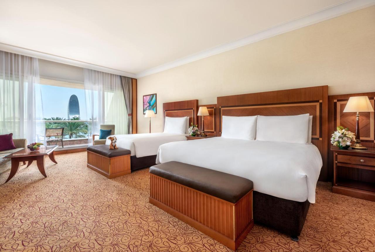 Al Raha Beach Resort & Spa Abu Dhabi Bagian luar foto The photo shows a spacious hotel room featuring two large beds with white linens and dark brown upholstered headboards. There are matching benches at the foot of each bed. The room is decorated with a warm color palette and has a carpeted floor. To o