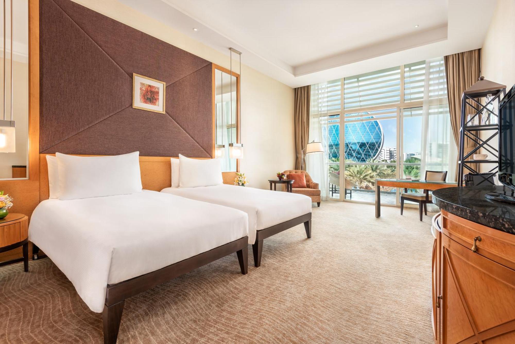 Al Raha Beach Resort & Spa Abu Dhabi Bagian luar foto The photo shows a well-decorated hotel room featuring two single beds with white linens. The room has a modern aesthetic, with a large window letting in natural light and offering a view of a circular structure outside, possibly a landmark or artisti