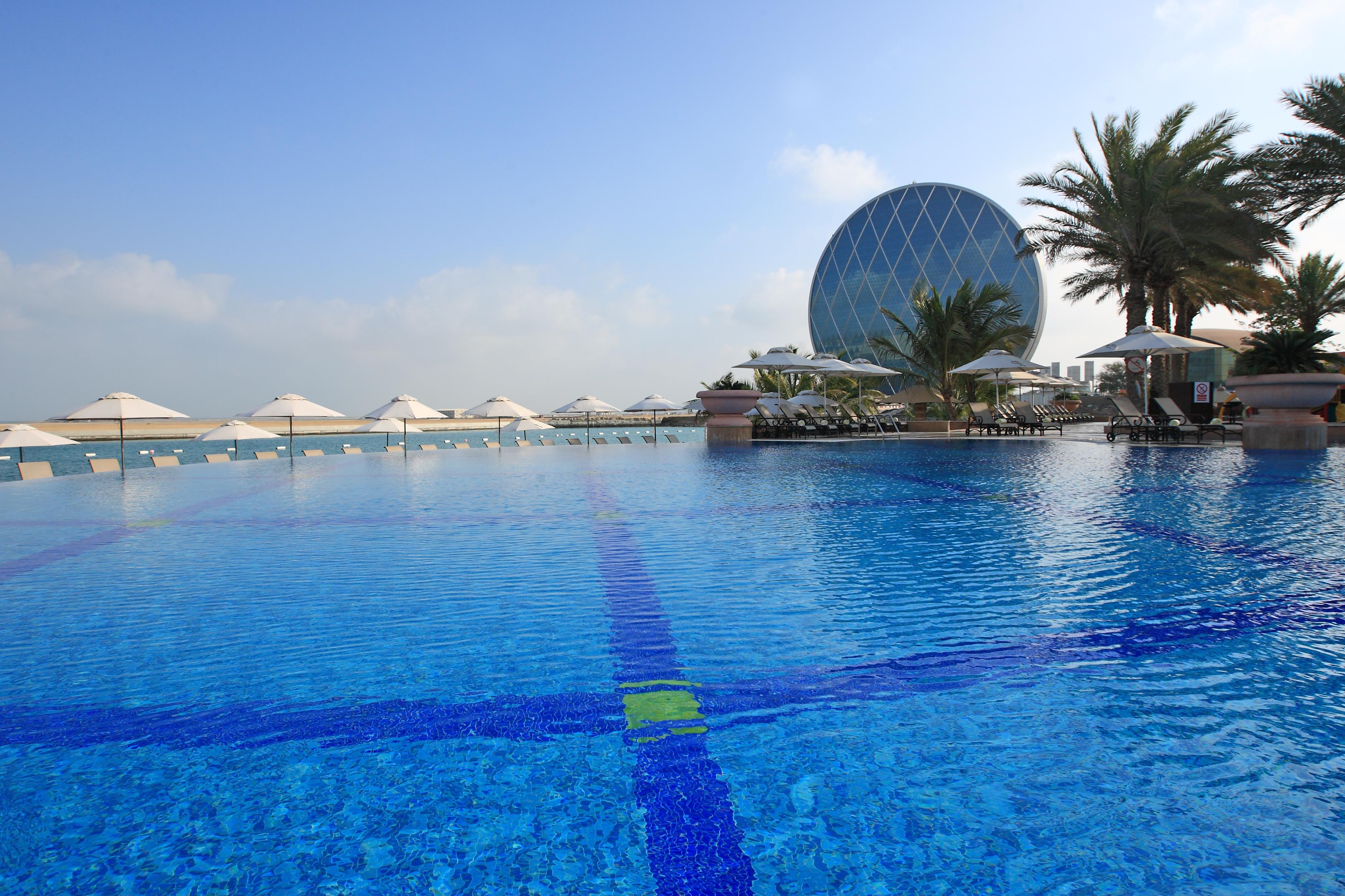 Al Raha Beach Resort & Spa Abu Dhabi Bagian luar foto The photo features a large, serene swimming pool with clear, blue water reflecting the sky. Surrounding the pool are several lounge chairs and umbrellas, suggesting a relaxed, resort-like atmosphere. In the background, a distinctive building with a r