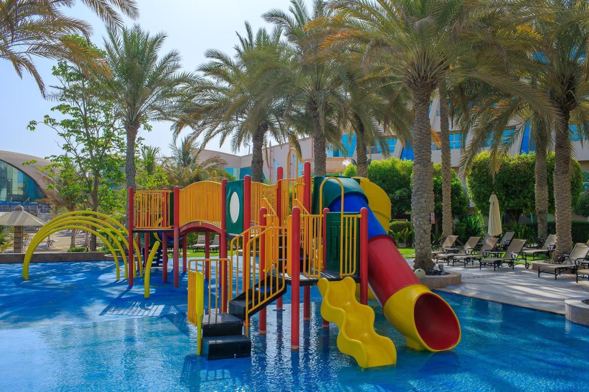 Al Raha Beach Resort & Spa Abu Dhabi Bagian luar foto The photo shows a colorful water play area located in a pool setting. It features a variety of play structures, including slides in multiple colors, such as yellow, red, and blue. Surrounding the play structure are palm trees, creating a tropical atm