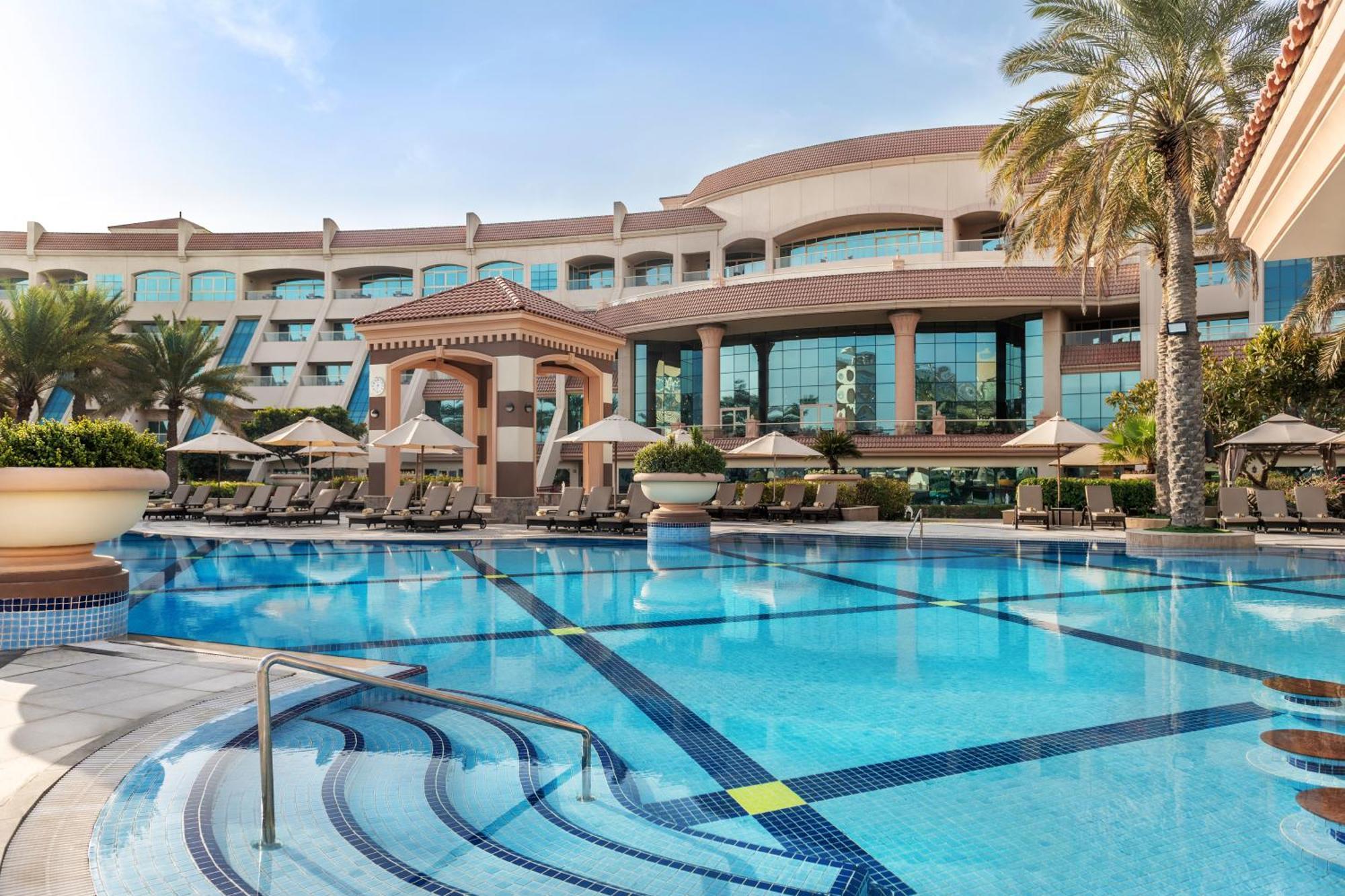Al Raha Beach Resort & Spa Abu Dhabi Bagian luar foto The photo shows a luxurious resort with a large swimming pool in the foreground. The pool has a clean, blue surface and features lounge chairs on the surrounding deck. In the background, there is a stylish and modern hotel building with multiple stor