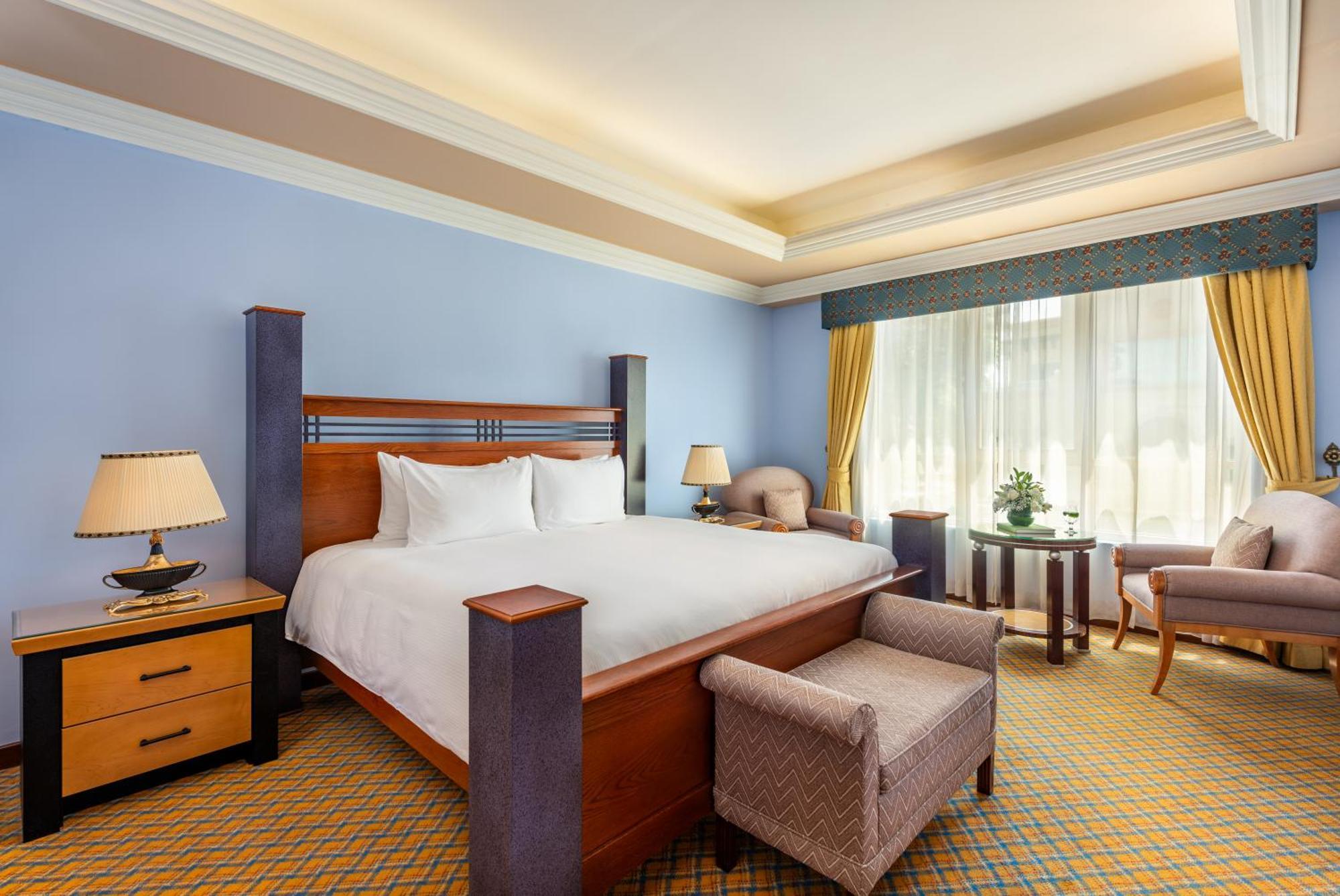 Al Raha Beach Resort & Spa Abu Dhabi Bagian luar foto The photo shows a well-decorated hotel room. It features a large bed with a wooden frame, neatly made with white bedding. On either side of the bed are bedside tables with lamps. There is a comfortable chair next to a window that has sheer curtains a