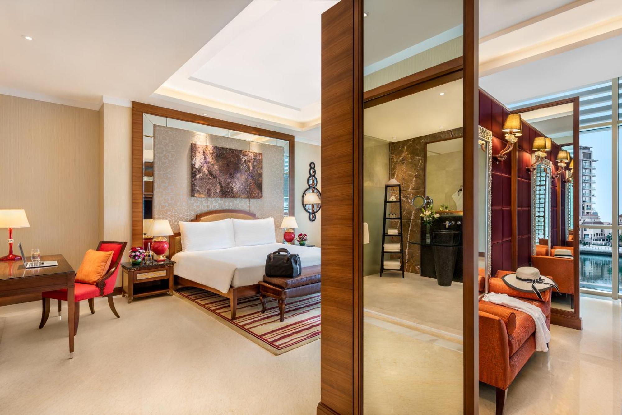 Al Raha Beach Resort & Spa Abu Dhabi Bagian luar foto The photo shows a luxurious hotel room with a modern design. The room features a large bed with white linens, flanked by bedside tables with lamps. There is a area with a chair and a small table on the left side, and a vibrant artwork hanging above t