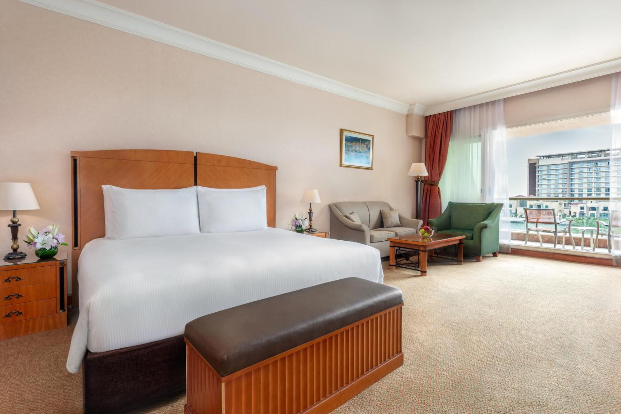 Al Raha Beach Resort & Spa Abu Dhabi Bagian luar foto The photo shows a well-furnished hotel room featuring a large bed with white linens and a wooden headboard. There's a brown bench at the foot of the bed. To one side, a sitting area includes a sofa and a coffee table, complemented by two lamps. Large