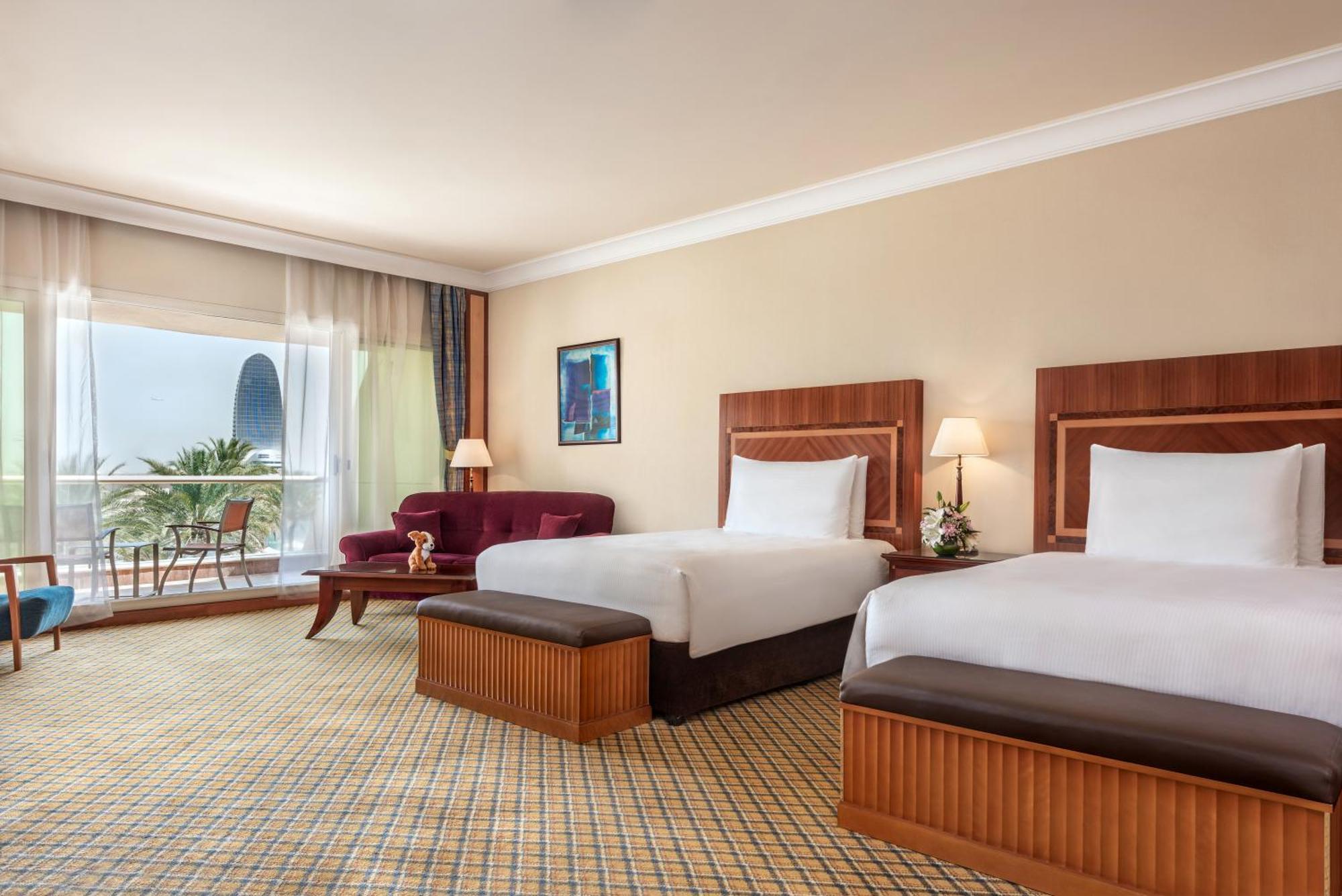 Al Raha Beach Resort & Spa Abu Dhabi Ruang foto The photo shows a hotel room featuring two twin beds with wooden headboards. The room has a light, neutral color scheme, with a large window that provides natural light and a view of the outdoors. There is a small seating area with a purple sofa and 