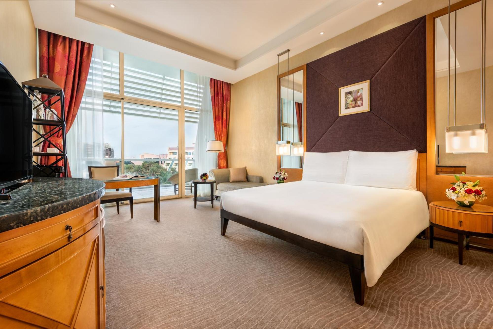 Al Raha Beach Resort & Spa Abu Dhabi Ruang foto The photo shows a modern hotel room featuring a large bed with a white duvet positioned against a purple accent wall. There are tall windows with sheer curtains allowing natural light to enter the space, along with red drapes framing the windows. A d