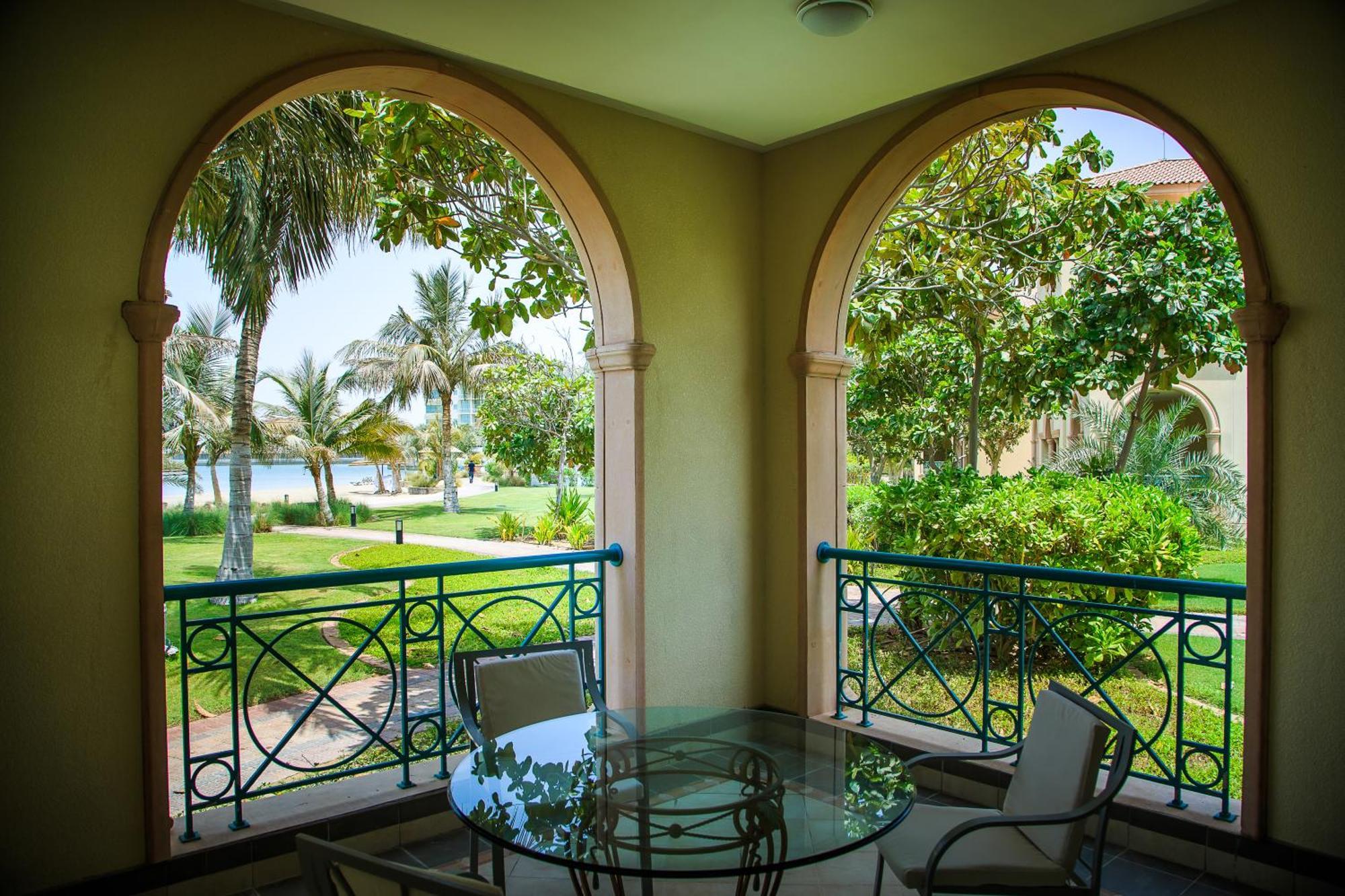 Al Raha Beach Resort & Spa Abu Dhabi Ruang foto The photo displays a beautifully designed balcony or terrace area. It features two large arches that frame the view outside. Through the arches, you can see a lush garden with palm trees and well-maintained greenery. There’s a glass table with severa