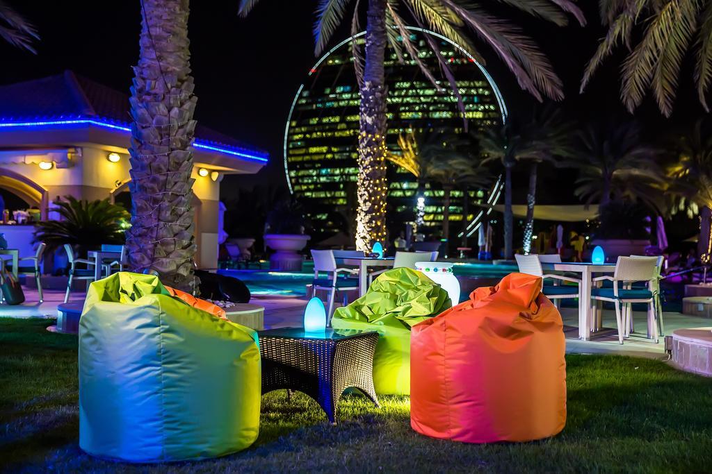 Al Raha Beach Resort & Spa Abu Dhabi Bagian luar foto The photo shows a vibrant outdoor lounge area at night. In the foreground, there are colorful bean bag chairs—one in bright green, one in yellow, and one in orange. A small table sits between the bean bags. In the background, illuminated palm trees a